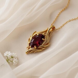 May include: A gold-colored pendant necklace featuring a large, oval-shaped, deep red gemstone. The gemstone is set in a decorative gold frame with a leaf-like design. The necklace chain is also gold.