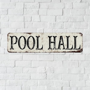 May include: A rectangular metal sign with a distressed, off-white finish and dark brown rust-like edges. The text POOL HALL is printed in a bold, black, serif font across the center. The sign features small circular holes at each of the four corners.