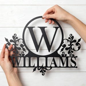 May include: A black metal wall sign featuring a large capital letter W centered within a circular frame. Below the circle, the name WILLIAMS is displayed in capital letters. Decorative scrollwork accents the sides and bottom of the sign.