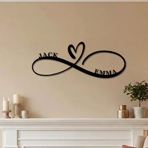 Puede incluir: Un letrero de pared de metal negro que presenta un símbolo de infinito entrelazado con una forma de corazón. El nombre JACK está integrado en el bucle izquierdo del símbolo de infinito y el nombre EMMA en el bucle derecho del símbolo.
