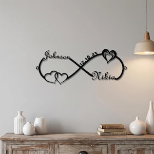 Puede incluir: Un letrero de pared de metal negro con forma de símbolo de infinito que presenta el nombre Johnson, el nombre Nikia, la fecha 12.16.23 y dos diseños de corazones. El letrero está montado en una pared e incluye pequeñas anillas circulares.