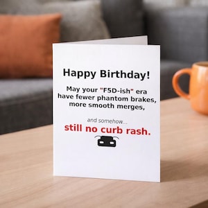 May include: A white greeting card with the text "Happy Birthday!" in black. The card includes humorous wishes for fewer phantom brakes and smooth merges, ending with "still no curb rash."