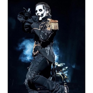 May include: A performer in a black military-style jacket with gold trim and epaulets, black trousers, and white face paint. The performer is on stage with a drum kit in the background, striking a pose with one hand raised.