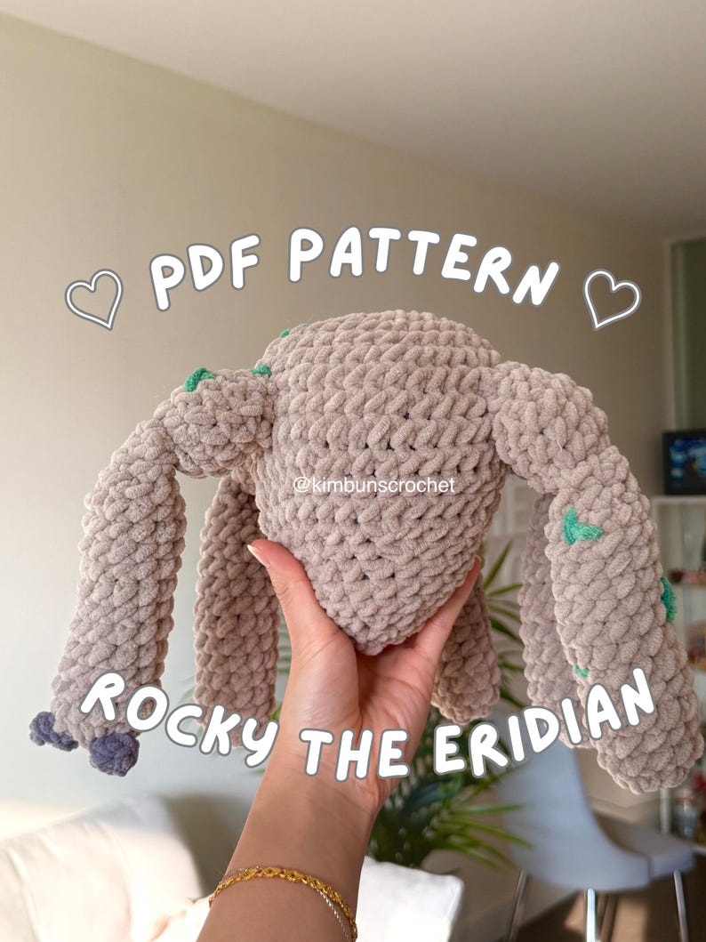 May include: A beige crocheted plush toy with four limbs and a rounded body. The toy has small green accents and is held in a person's hand. Text on the image reads "PDF PATTERN" and "ROCKY THE ERIDIAN".