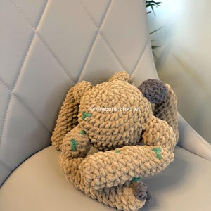 May include: A handmade crochet bunny in shades of tan and gray, with green accents. The plush toy is made of soft, textured yarn and is positioned on a light gray chair. The bunny's ears and limbs are visible, showcasing the detailed crochet work.