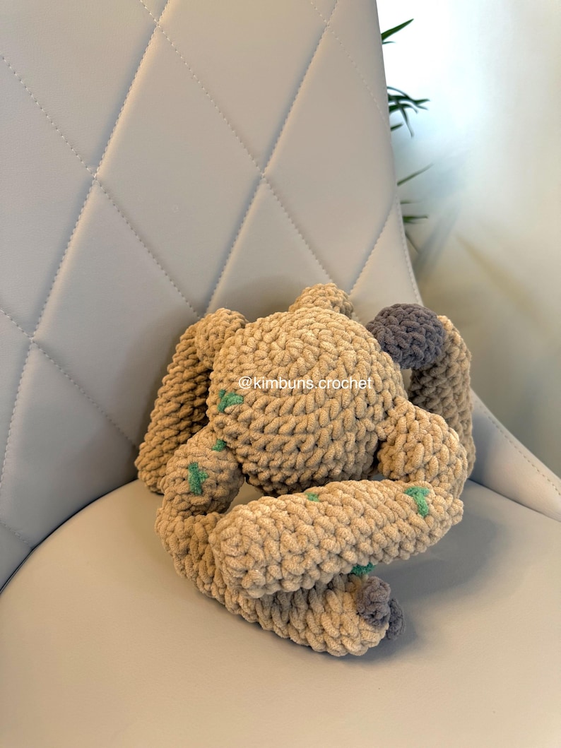 May include: A handmade crochet bunny in shades of tan and gray, with green accents. The plush toy is made of soft, textured yarn and is positioned on a light gray chair. The bunny's ears and limbs are visible, showcasing the detailed crochet work.