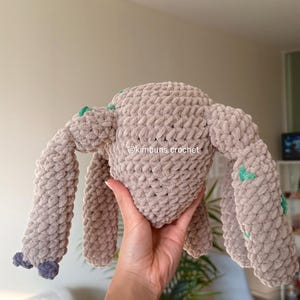 May include: A handmade, beige crochet creature with four extended limbs and a rounded head. The plush toy has small green accents and a purple detail on the feet. The text "@kimbuns.crochet" is visible on the toy.