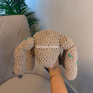 May include: A handmade, beige crochet bunny plushie with long ears and four limbs. The plushie has small green accents and a gray pom-pom tail. The text "@kimbuns.crochet" is visible on the plushie.