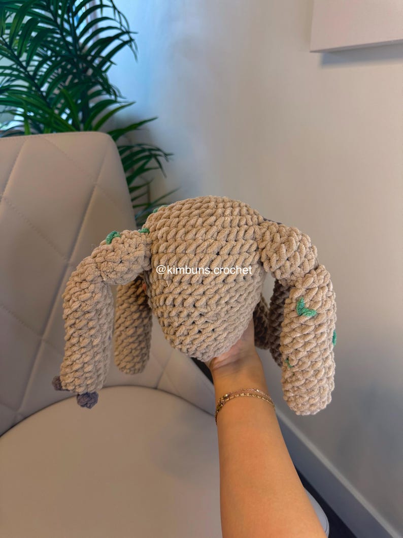 May include: A handmade, beige crochet bunny plushie with long ears and four limbs. The plushie has small green accents and a gray pom-pom tail. The text "@kimbuns.crochet" is visible on the plushie.