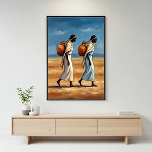 May include: A framed wall art print depicting two figures walking across a desert landscape, each carrying a large orange clay water vessel on their back. The figures wear simple, flowing robes in white and light blue against a deep blue sky.
