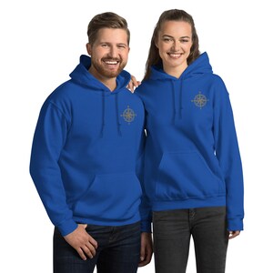 Puede incluir: Dos personas con sudaderas con capucha azul real a juego. Cada sudadera tiene un bolsillo delantero y un diseño de brújula dorada bordado en el pecho. Las sudaderas están hechas de un material suave.