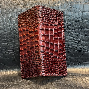 May include: A rectangular, red-brown leather wallet with a crocodile skin texture. The wallet is upright, displaying its detailed pattern and colour variations. The background features a similar textured surface.