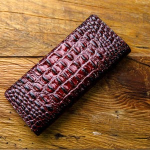 May include: A burgundy leather wallet with a crocodile skin texture. The rectangular wallet is glossy and reflects light, with a pattern of raised, rounded scales. The wallet is lying on a wooden surface.