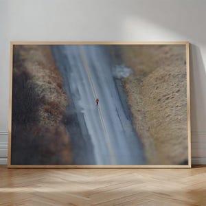 May include: A framed landscape photograph featuring an aerial view of a lone runner in an orange shirt jogging along a grey asphalt road with double yellow lines, flanked by dry, brownish-yellow grassy hillsides and dark, muted vegetation.