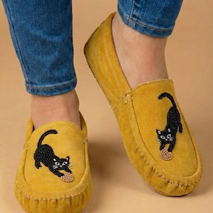 May include: Mustard yellow suede moccasins with a black beaded cat design. The cat is playing with a small ball. The shoes have a rounded toe and a stitched sole. The person is wearing blue jeans.