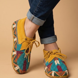 Könnte beinhalten: Senfgelbe Mokassin-Schuhe mit aufwendigen Perlenarbeiten in türkisfarbenen, roten und orangefarbenen geometrischen Mustern. Die Schuhe haben ein Obermaterial aus Wildleder und Schnürsenkel. Die Person trägt eine blaue Jeans.