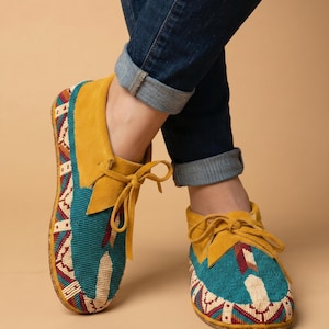 May include: Moccasin-style shoes with a teal beaded vamp and a mustard yellow suede collar and tongue. The shoes have a tan sole and are tied with mustard yellow laces. The shoes are worn with blue jeans.