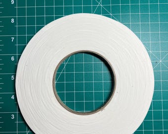 May include: A roll of white double-sided tape with a brown cardboard core. The tape is on a green cutting mat with inch measurements along the bottom and side.