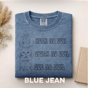 May include: A blue jean colored t-shirt with a Comfort Colors label at the neck. The graphic features three stacked cats and the text: HEAR NO EVIL, SPEAK NO EVIL, SEE NO EVIL. The top cat covers its ears, the middle cat covers its mouth, and the bottom cat covers its eyes.
