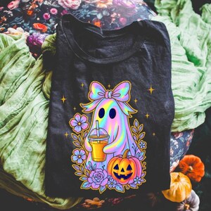 May include: A black t-shirt featuring a graphic of a holographic ghost wearing a large bow, holding an iced beverage, and standing next to a jack-o'-lantern surrounded by purple and blue flowers and small yellow stars against a dark background.