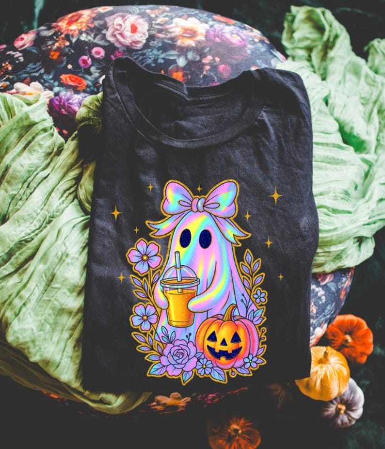 May include: A black t-shirt featuring a graphic of a holographic ghost wearing a large bow, holding an iced beverage, and standing next to a jack-o'-lantern surrounded by purple and blue flowers and small yellow stars against a dark background.