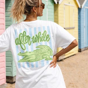 May include: White t-shirt with a green alligator graphic and the words "after while" in green script. The design is on the back of the shirt, with blue and white vertical stripes. The person is wearing sunglasses and bracelets.