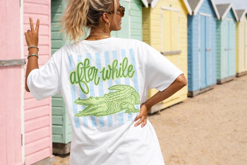 May include: White t-shirt with a green alligator graphic and the words "after while" in green script. The design is on the back of the shirt, with blue and white vertical stripes. The person is wearing sunglasses and bracelets.