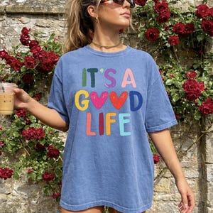 May include: Blue short-sleeved t-shirt with the text "IT'S A GOOD LIFE" in colorful letters. The shirt has a vintage wash appearance. The image is set against a backdrop of red roses and a stone wall.