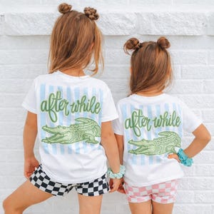 May include: Two children wearing white t-shirts with the text "after while" and a green alligator graphic. The shirts have blue striped accents. One child wears black and white checkered shorts, the other wears pink and white checkered shorts.