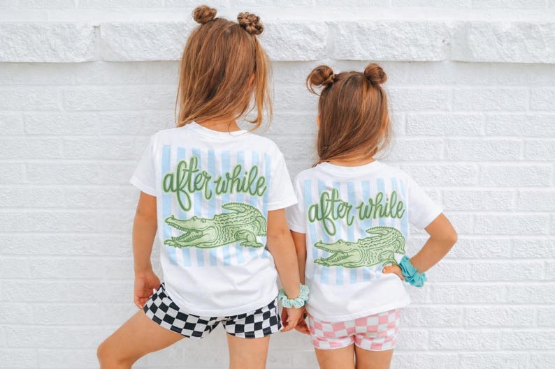May include: Two children wearing white t-shirts with the text "after while" and a green alligator graphic. The shirts have blue striped accents. One child wears black and white checkered shorts, the other wears pink and white checkered shorts.