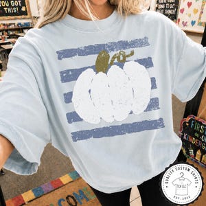 May include: A light blue short-sleeve t-shirt featuring a large, white, distressed-texture pumpkin graphic centered over horizontal, faded navy blue stripes. The pumpkin has a textured olive green stem and a curled vine detail at the top.