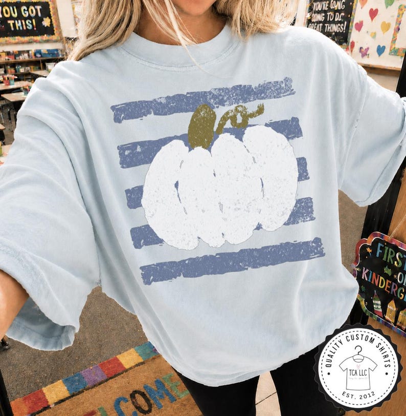 May include: A light blue short-sleeve t-shirt featuring a large, white, distressed-texture pumpkin graphic centered over horizontal, faded navy blue stripes. The pumpkin has a textured olive green stem and a curled vine detail at the top.
