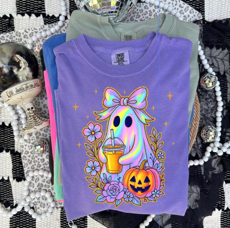 May include: A purple t-shirt featuring a graphic of a holographic ghost wearing a large bow, holding an iced coffee, and sitting next to a jack-o'-lantern surrounded by flowers and stars. The shirt has a Comfort Colors brand label at the neck.