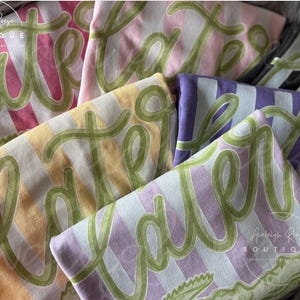 May include: Folded t-shirts in various pastel colors, including pink, yellow, and purple, with a striped pattern. Each shirt features the word "Easter" in green script. The shirts are from the Analeigh Reese Boutique.
