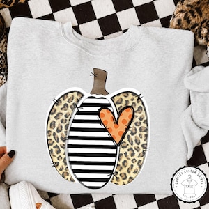 May include: A light gray crewneck sweatshirt featuring a central graphic of a pumpkin. The pumpkin design consists of black and white horizontal stripes, leopard print sections, and an orange heart with polka dots, accented by black stitch marks.