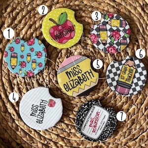 May include: A collection of seven round car coasters with various teacher-themed designs. The coasters feature images of pencils, apples, and composition books, with text such as "Principal", "Miss Elizabeth", and "1st Grade Teacher".