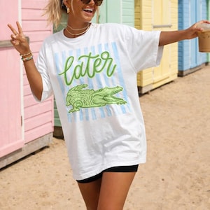 May include: White t-shirt with a green alligator graphic and the word "Later" in green script. The shirt has blue and white stripes behind the alligator. The person is wearing black shorts and sunglasses.