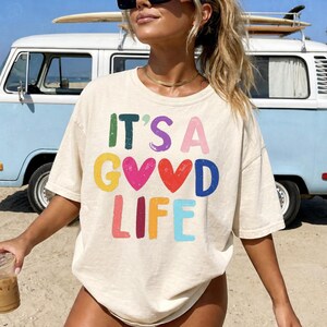 May include: Cream-colored t-shirt with the phrase "IT'S A GOOD LIFE" in colorful, distressed lettering. The shirt is short-sleeved and appears to be made of a soft material. A light blue van is in the background.