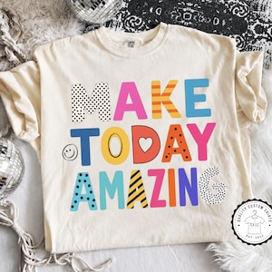 May include: A cream-colored short-sleeve t-shirt featuring the multi-colored text MAKE TODAY AMAZING in various patterns including polka dots, stripes, and solid colors. The letter O in TODAY is a heart, and a small smiley face is on the left.