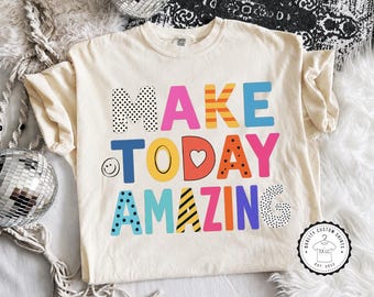 May include: A cream-colored short-sleeve t-shirt featuring the multi-colored text MAKE TODAY AMAZING in various patterns including polka dots, stripes, and solid colors. The letter O in TODAY is a heart, and a small smiley face is on the left.