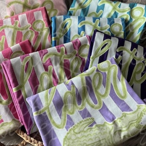 May include: Folded t-shirts in various colors, including pink, blue, and purple, with white stripes. Each shirt features the word "Later" in a green, cursive font and a green alligator graphic.