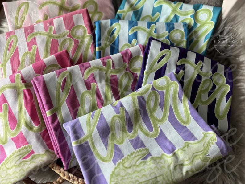 May include: Folded t-shirts in various colors, including pink, blue, and purple, with white stripes. Each shirt features the word "Later" in a green, cursive font and a green alligator graphic.