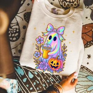 May include: A beige crewneck sweatshirt featuring a graphic print of a holographic ghost wearing a large bow, holding an iced beverage, and standing next to a jack-o'-lantern surrounded by purple and blue flowers with small yellow stars.