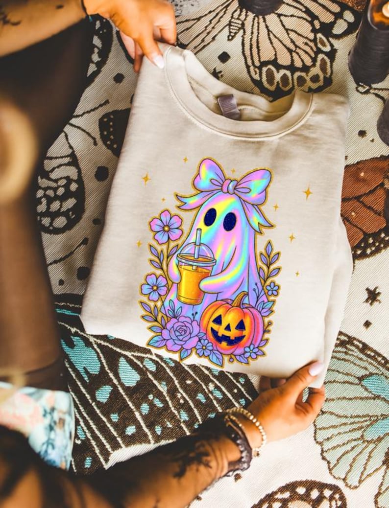 May include: A beige crewneck sweatshirt featuring a graphic print of a holographic ghost wearing a large bow, holding an iced beverage, and standing next to a jack-o'-lantern surrounded by purple and blue flowers with small yellow stars.