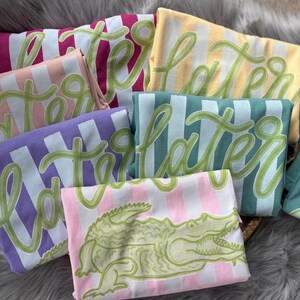May include: A collection of folded t-shirts in various colors, including pink, purple, yellow, and teal. Each shirt features the word "Later" in a green script font over vertical stripes. One shirt displays a green alligator graphic.