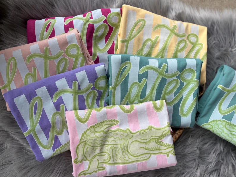 May include: A collection of folded t-shirts in various colors, including pink, purple, yellow, and teal. Each shirt features the word "Later" in a green script font over vertical stripes. One shirt displays a green alligator graphic.