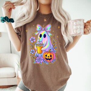 May include: A brown t-shirt featuring a graphic of a holographic ghost wearing a bow, holding an iced coffee, and surrounded by purple flowers and a jack-o'-lantern pumpkin. The design includes small yellow star accents around the central ghost figure.