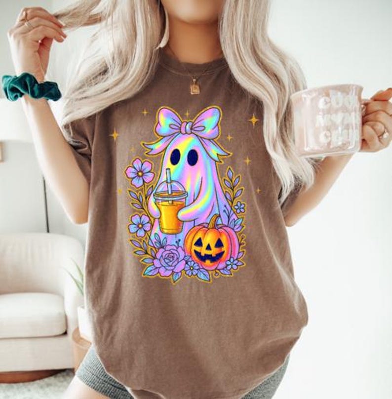May include: A brown t-shirt featuring a graphic of a holographic ghost wearing a bow, holding an iced coffee, and surrounded by purple flowers and a jack-o'-lantern pumpkin. The design includes small yellow star accents around the central ghost figure.