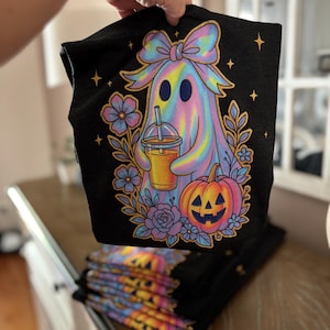 May include: A black t-shirt featuring a graphic of a holographic-style ghost wearing a large bow, holding an iced beverage, and standing next to a jack-o'-lantern. The design is surrounded by floral elements and small four-pointed stars in shades of pink, blue, and orange.