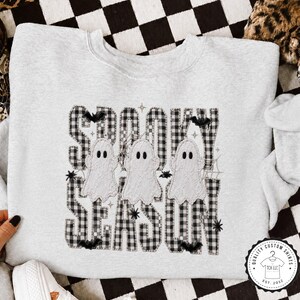 May include: A light gray crewneck sweatshirt featuring a graphic with the text SPOOKY SEASON in a black and white buffalo plaid pattern. The design includes three white ghosts, small black bats, spider webs, and stars scattered throughout.